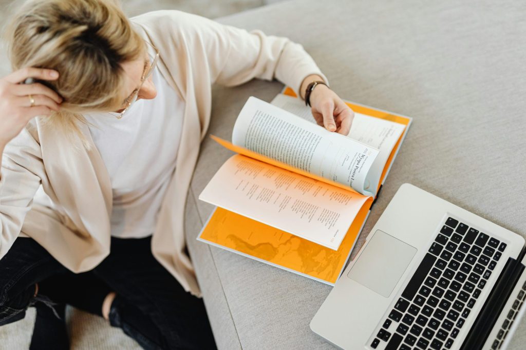 Woman sitting on a sofa, browsing a book with orange pages, laptop nearby, in a relaxed setting.