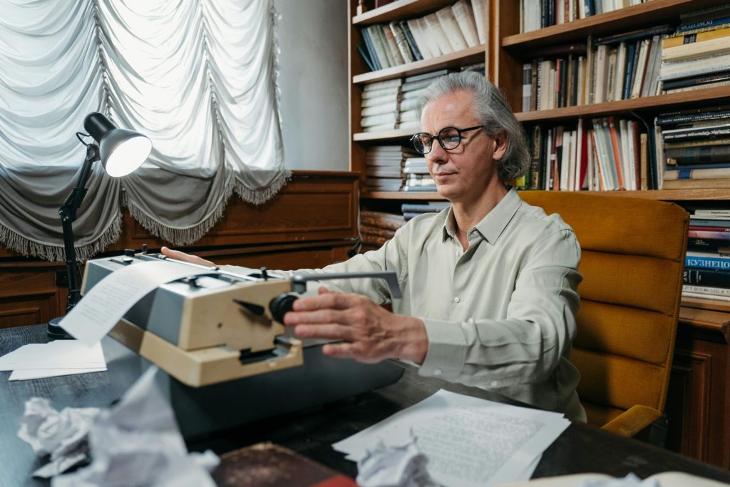Elderly man writing on a typewriter in a book-filled study, showcasing wisdom and creativity.